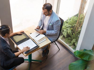 Above view of serious business partners having meeting in cafe. Latin American businessman and Asian businesswoman sitting together at table, man using mobile phone. Meeting concept